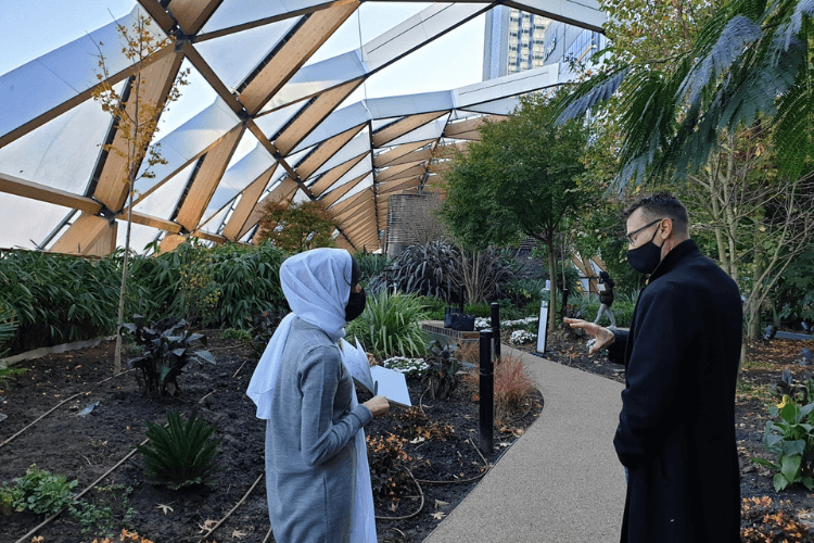 2 people talking in the middle of a garden sitting underneath a large architectural tunnel structure