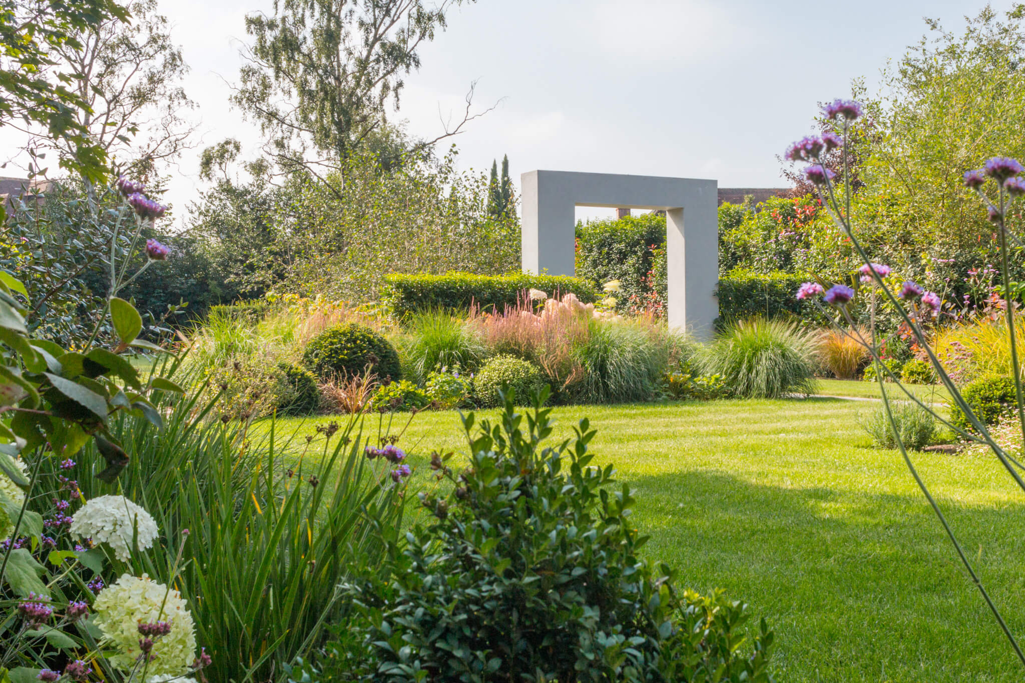 A square stone archway between two hedges leading to another section of a garden